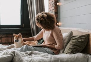 Black woman with afro hair reads on bed with her dog, cozy indoor setting.