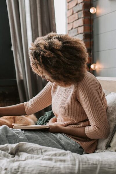 Black woman with afro hair reads on bed with her dog, cozy indoor setting.