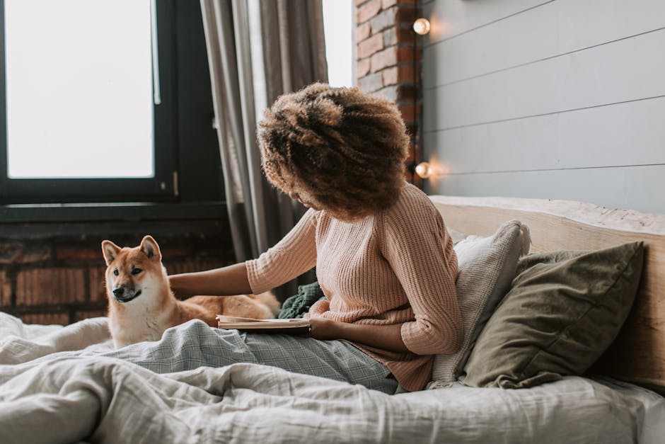 Black woman with afro hair reads on bed with her dog, cozy indoor setting.